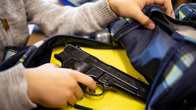 A boy's hands placing a black handgun inside a blue and green plaid backpack.